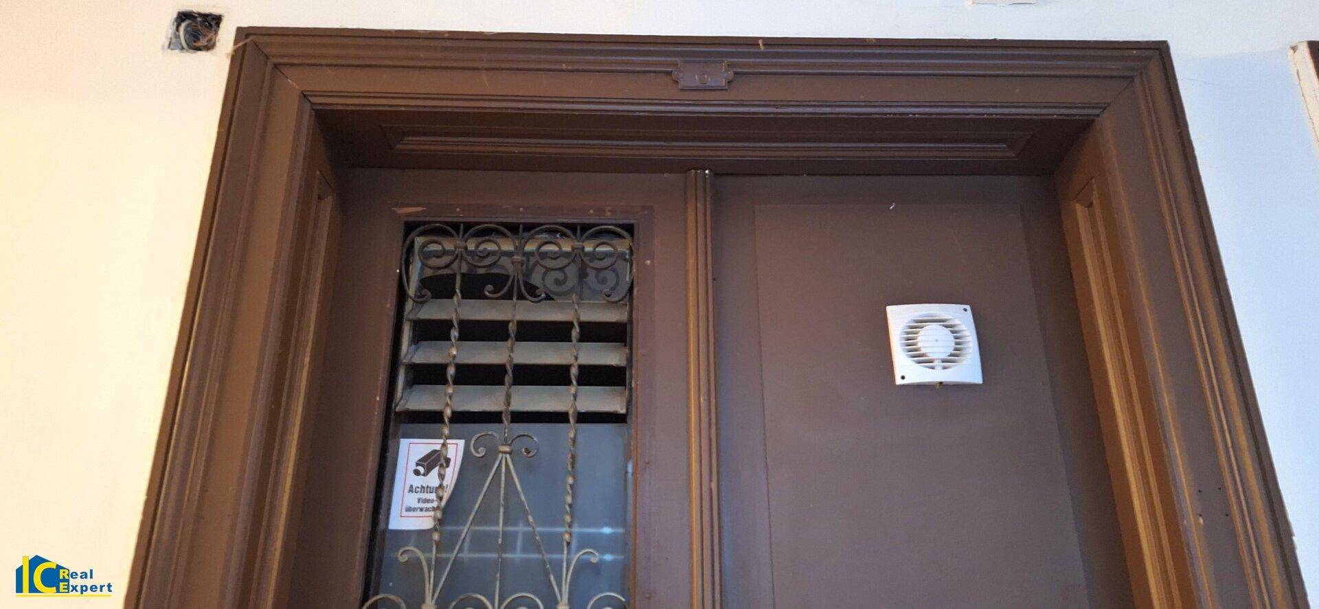 Alte Holztür mit Gitterfenster und einem kleinen Abluftventilator an der Wand.