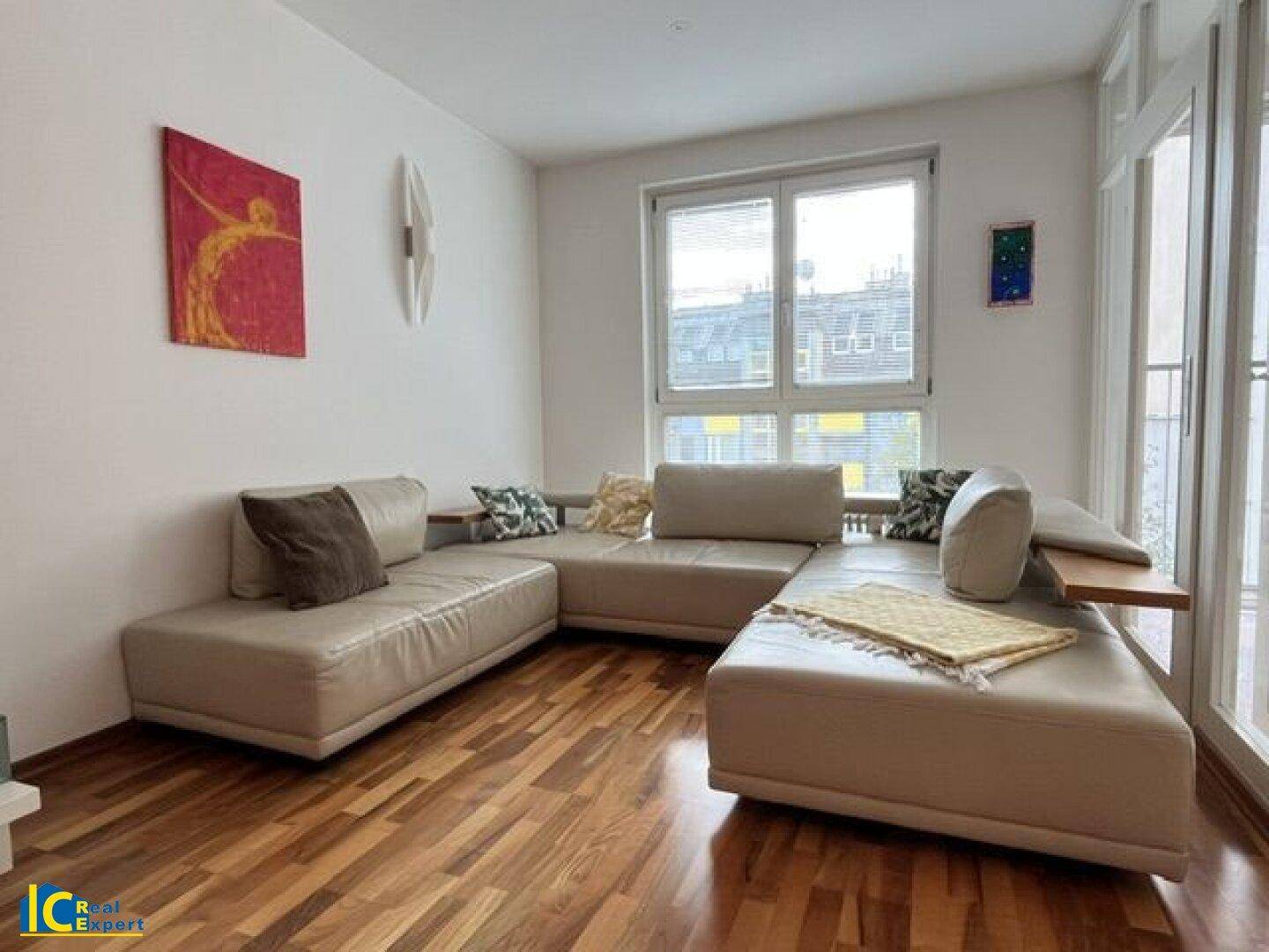 Gemütliches Wohnzimmer mit beigem Ecksofa, Holzboden und großen Fenstern für viel Tageslicht.