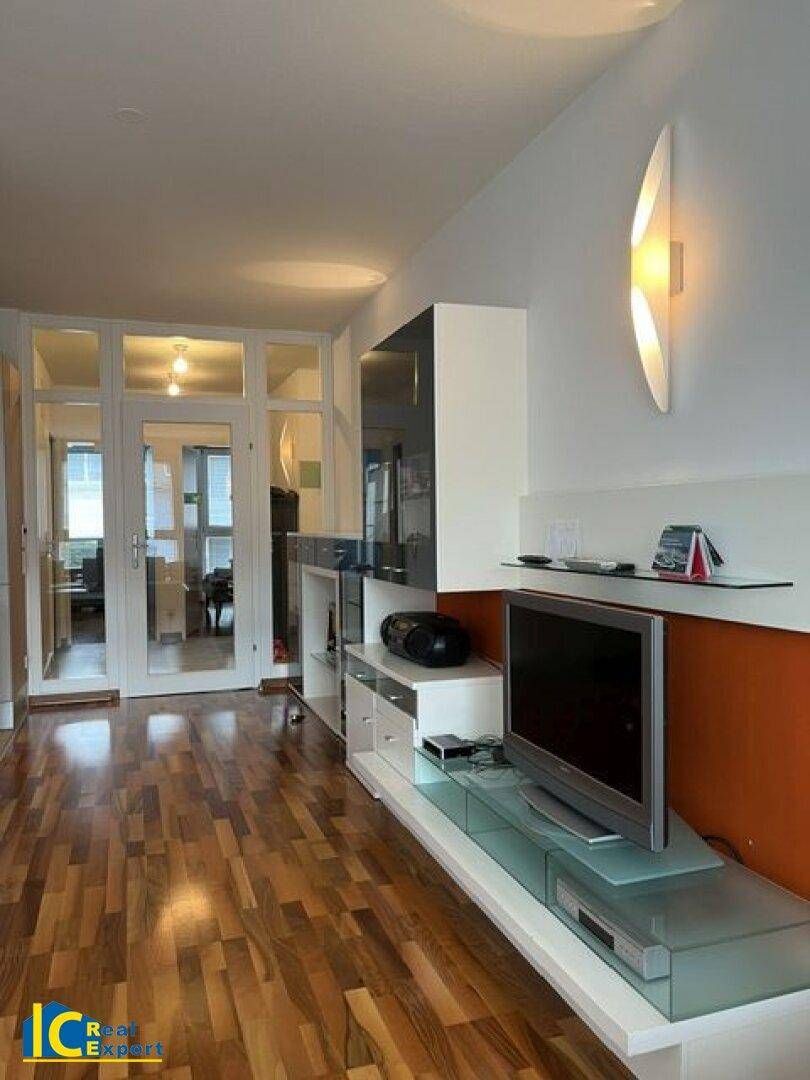 Wohnzimmer mit Holzboden, modernem TV-Möbel und Zugang zum verglasten Balkon.