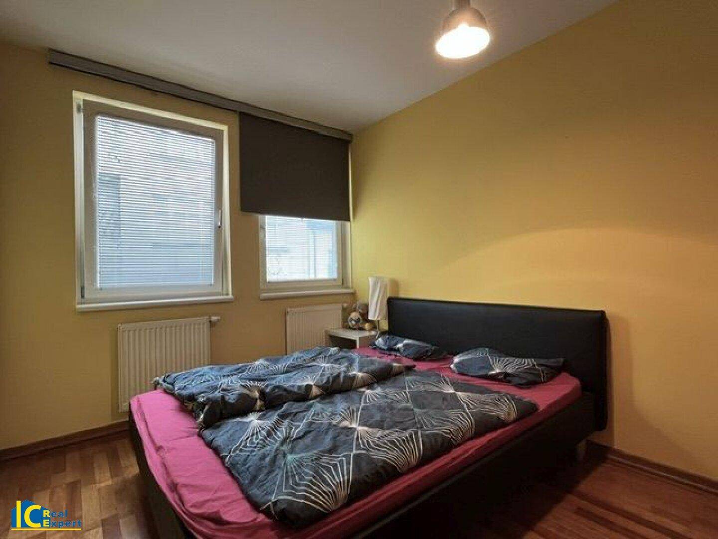 Schlafzimmer mit gelben Wänden, Doppelbett und zwei Fenstern für natürliche Belichtung.