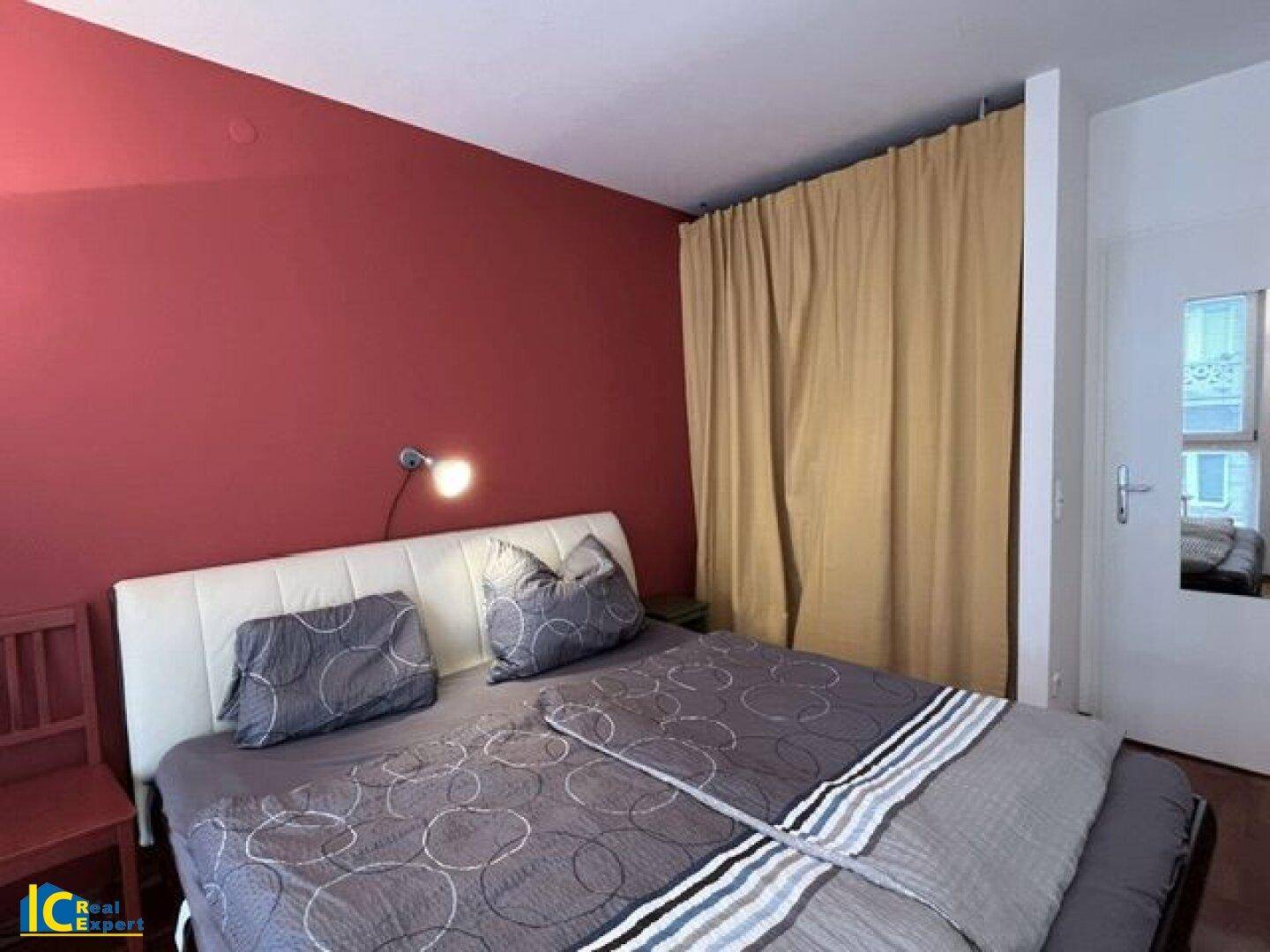 Schlafzimmer mit roter Akzentwand, Doppelbett und Vorhang, der einen Bereich abtrennt.