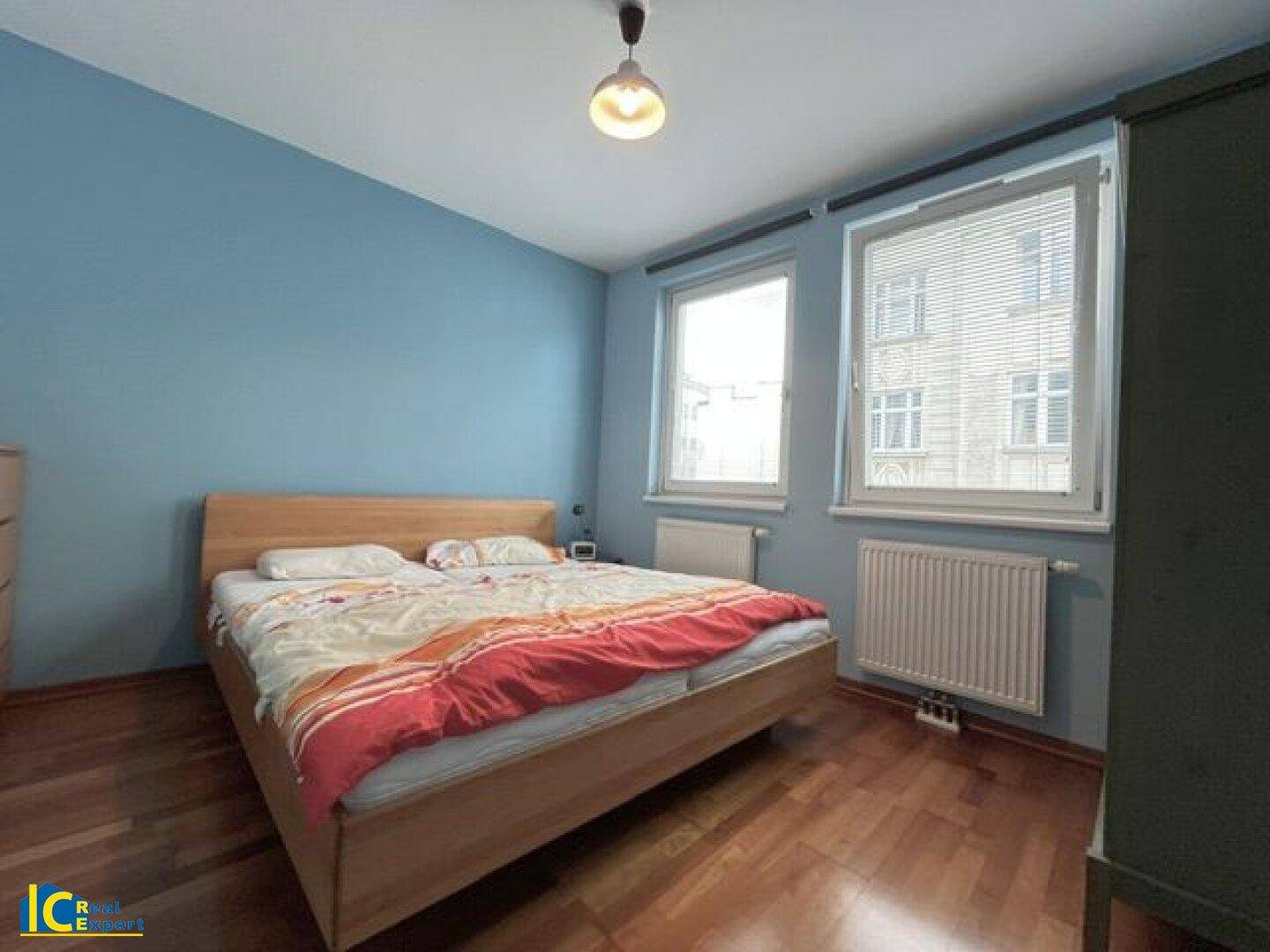 Schlafzimmer mit blauen Wänden, Holzbett und zwei Fenstern für helle Atmosphäre.