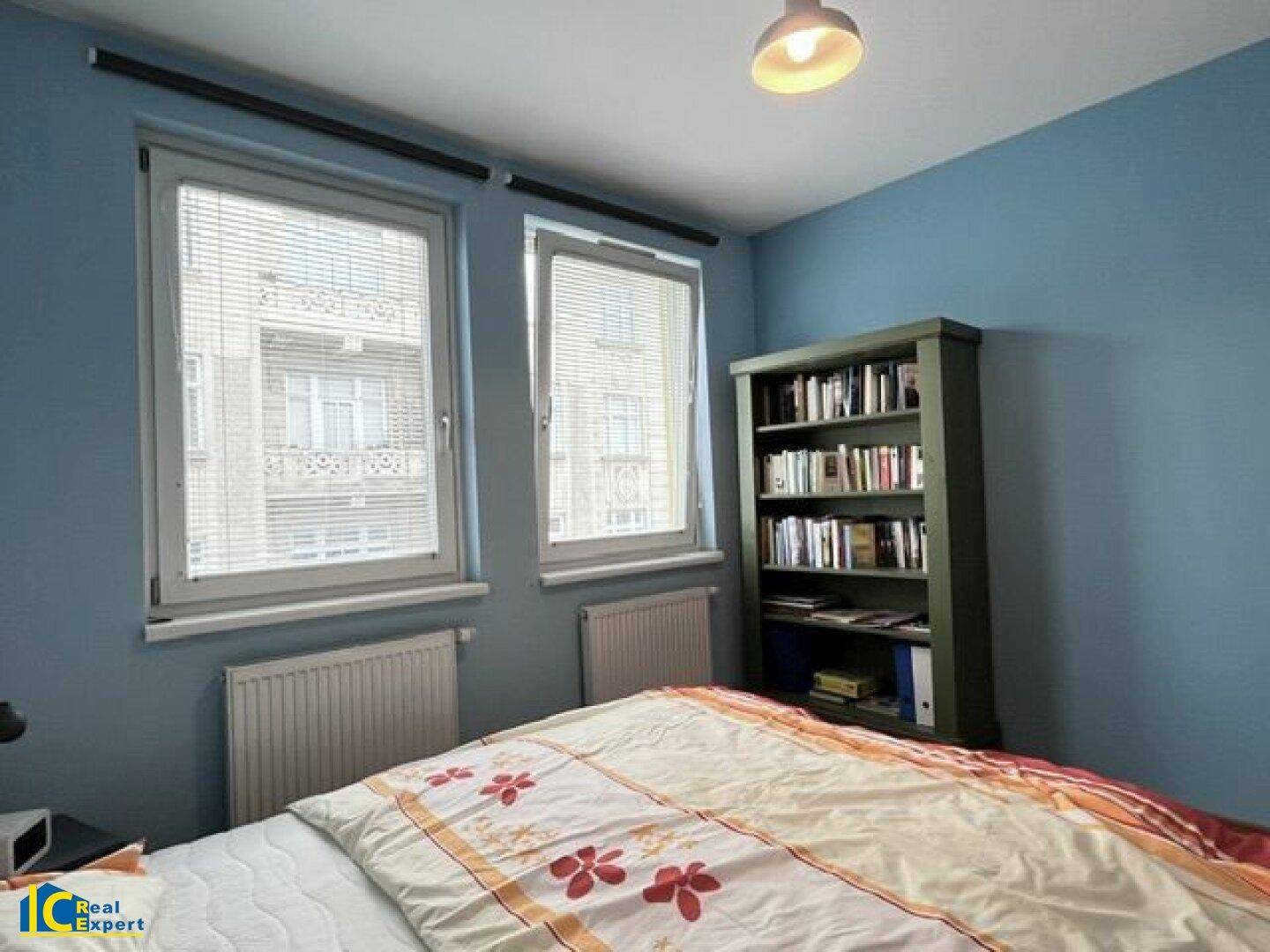 Schlafzimmer mit blauen Wänden, Holzbett und Bücherregal neben den Fenstern.