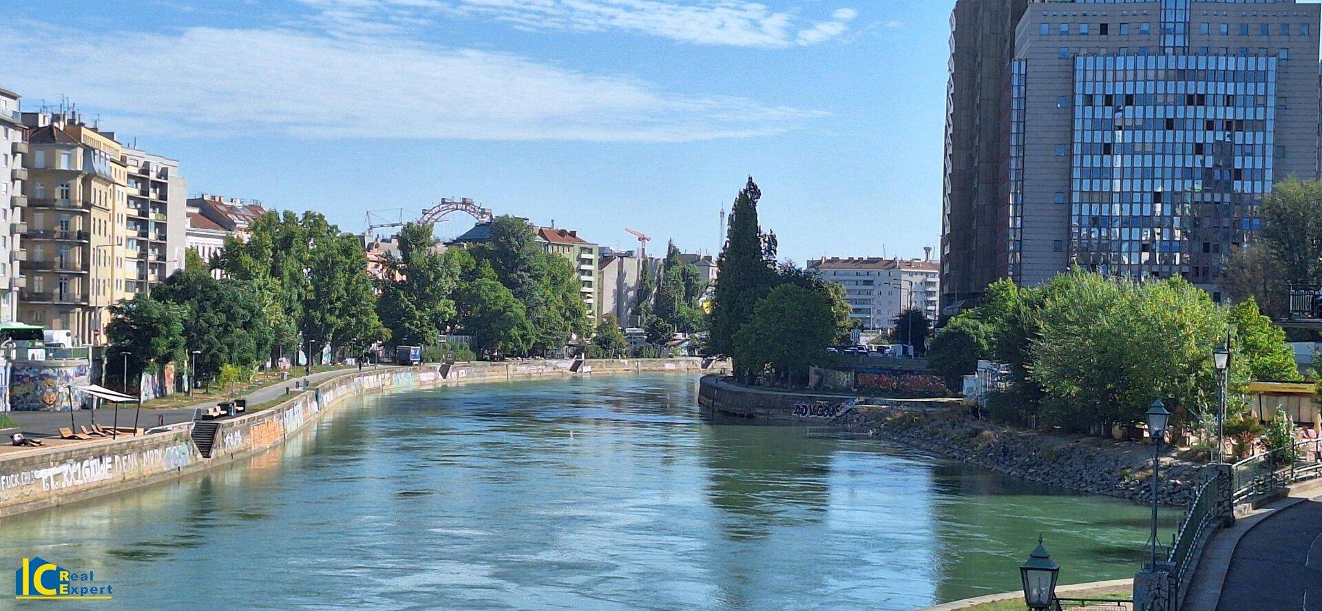 Panoramablick auf einen Fluss oder Kanal mit Gebäuden, Bäumen und einem Riesenrad in der Ferne.