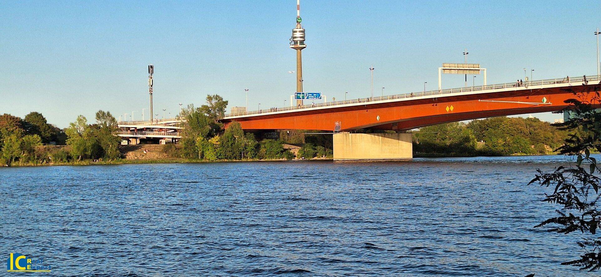 Weitläufiger Blick über die Donau mit einer markanten roten Brücke und dem Donauturm am Horizont.