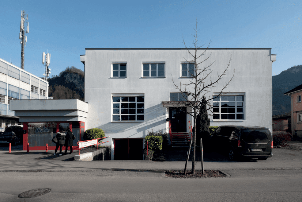 Moderne weiße Gebäudefassade mit großen Fenstern, rotem Eingangsbereich und geparktem Fahrzeug.