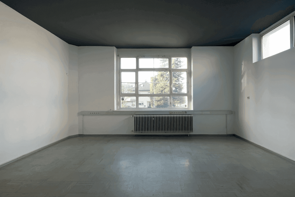 Leerer Raum mit weiß gestrichenen Wänden, dunkler Decke und einem großen Fenster mit Blick ins Grüne.