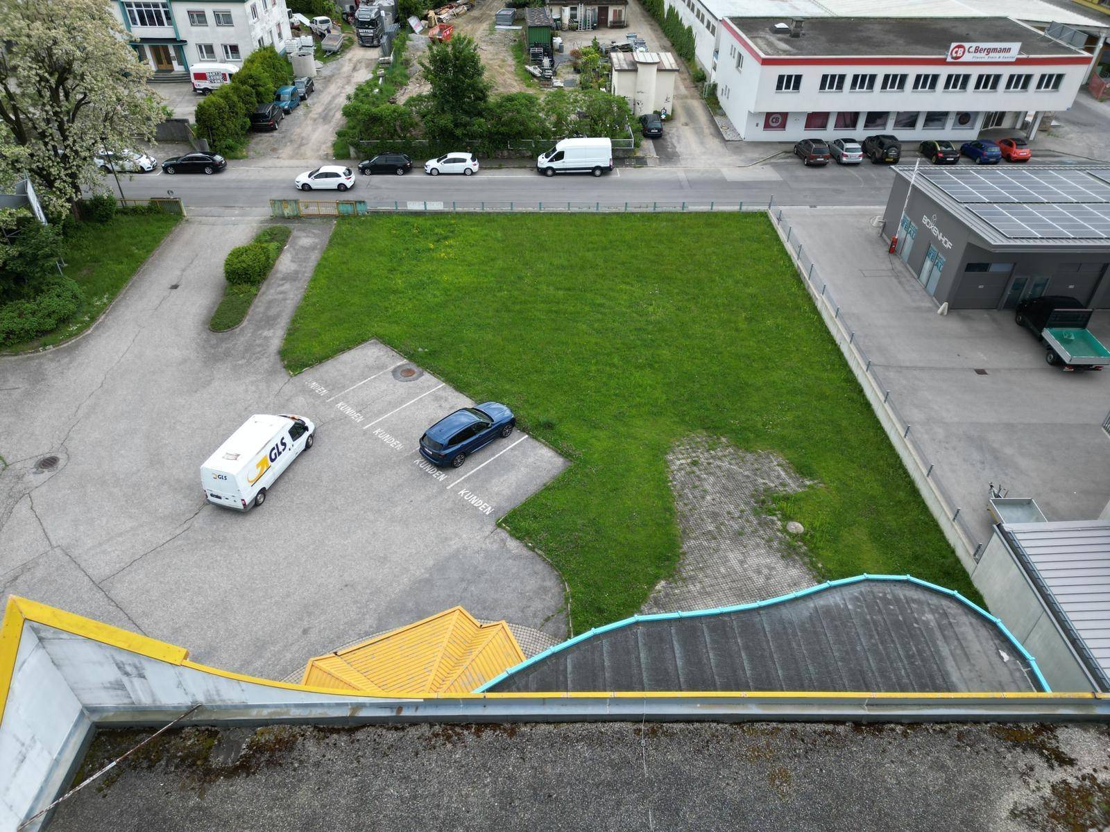 Luftaufnahme eines grünen Grundstücks mit Parkplätzen, umgeben von Geschäftsgebäuden und einer Straße.