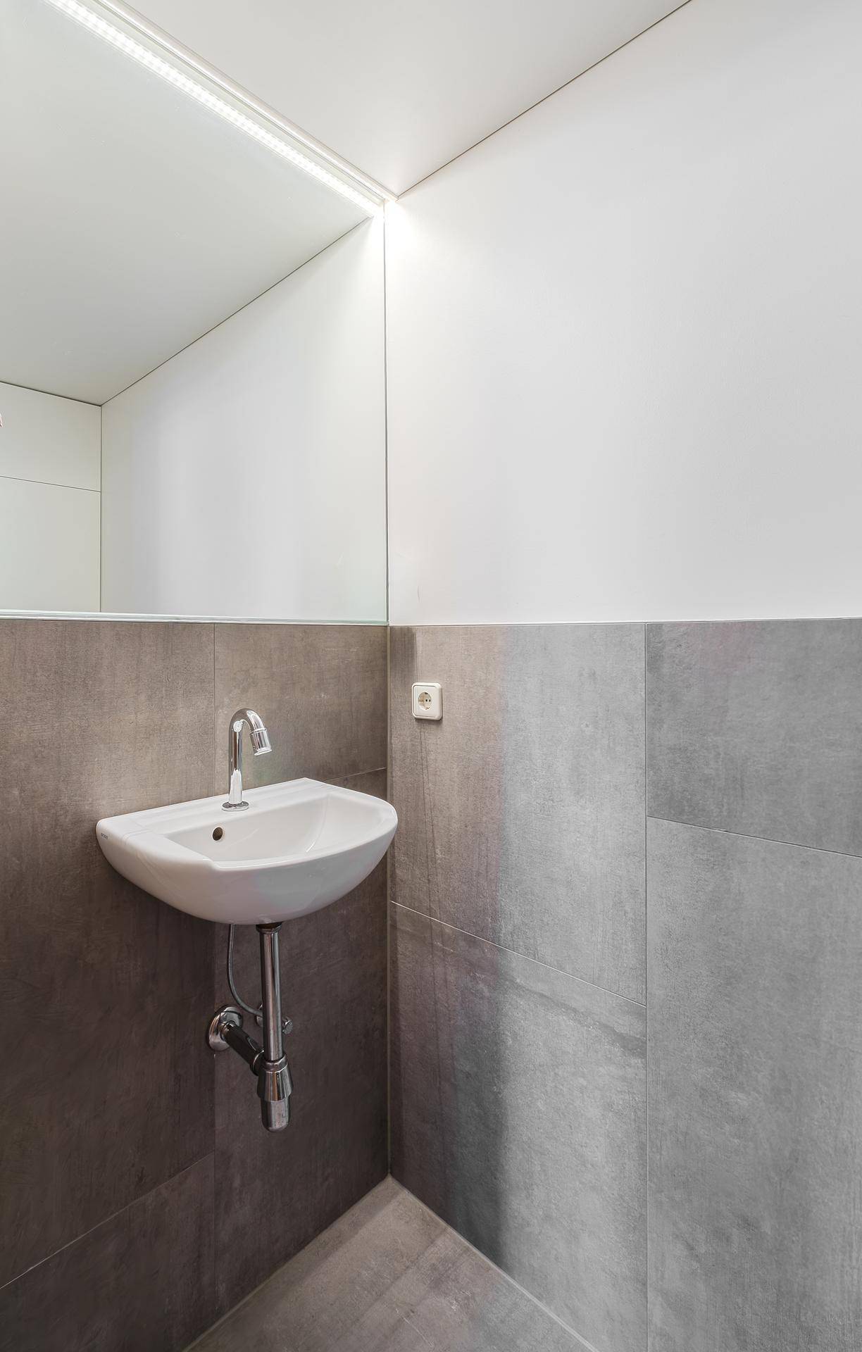 Modernes Gäste-WC mit Waschbecken, großem Spiegel und grauen Wandfliesen.
