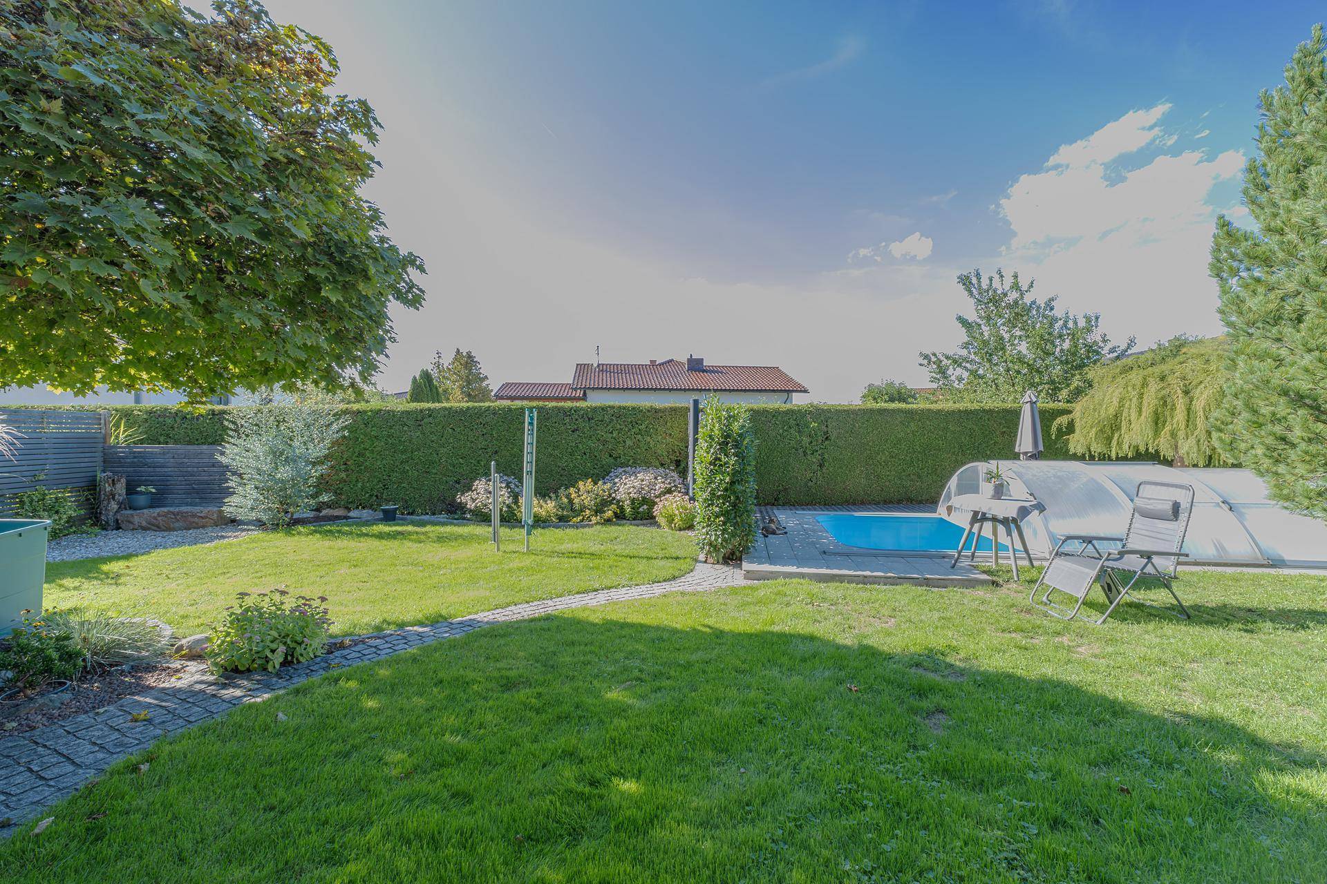Weitläufiger Garten mit großer Rasenfläche, hohen Hecken und dem überdachten Swimmingpool im Hintergrund.
