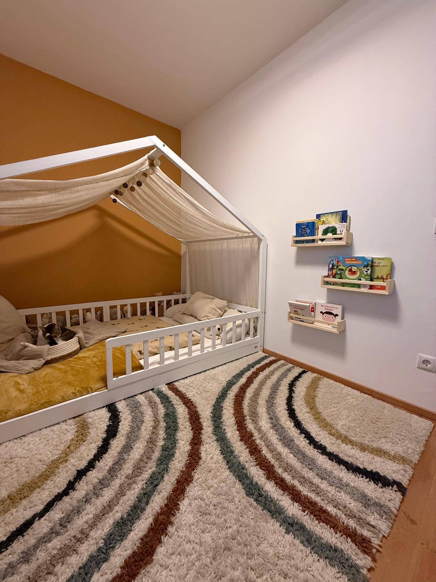 Kinderzimmer mit Himmelbett, farbenfrohem Teppich und Bücherregalen an der Wand.