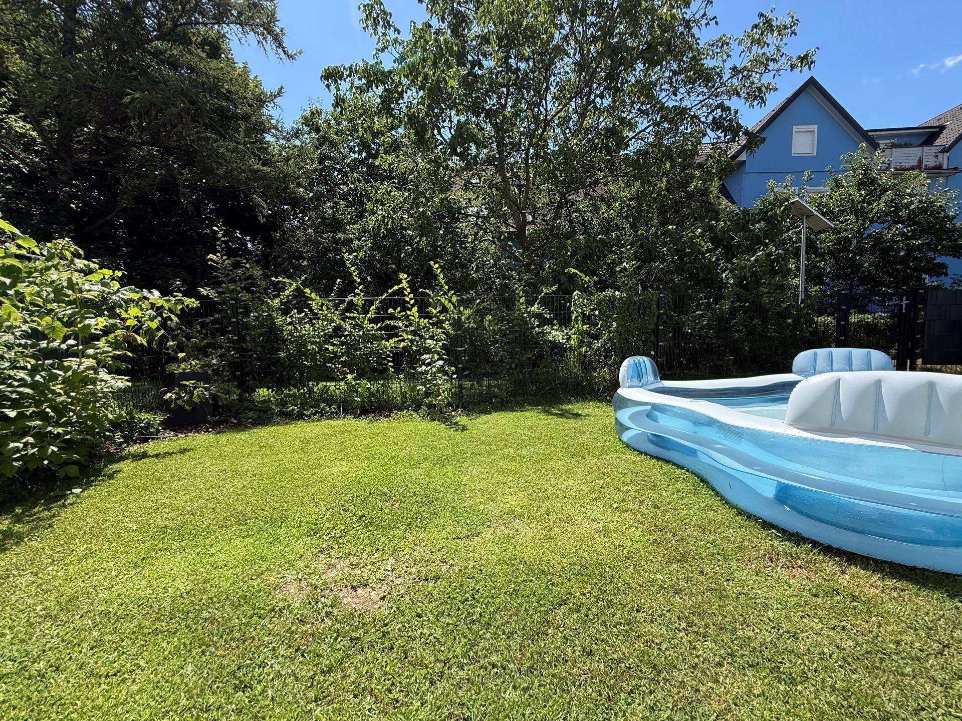 Grüner Garten mit einem aufblasbaren blauen Pool auf der Rasenfläche, umgeben von Bäumen und einem Haus.