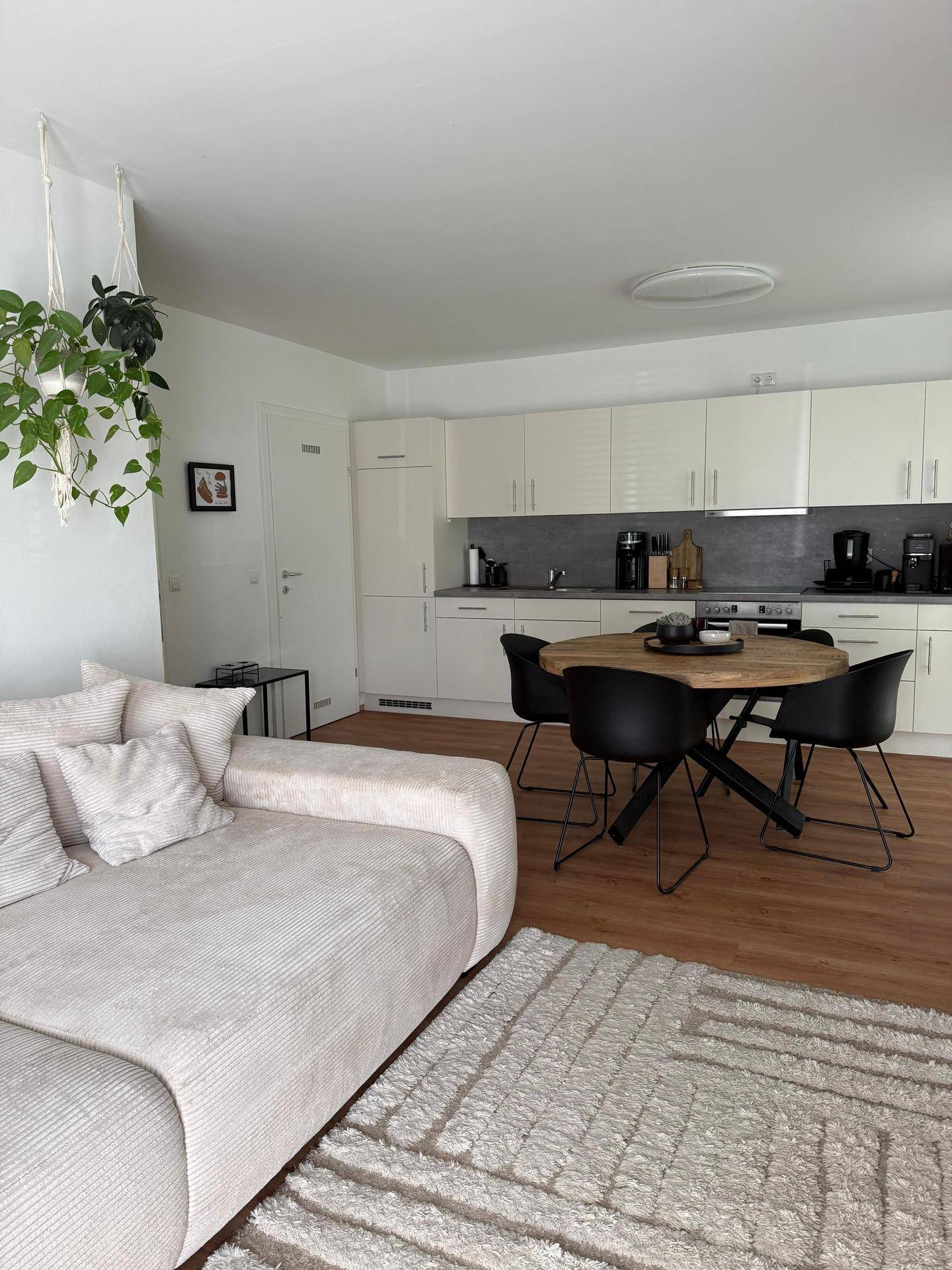 Offener Wohn-Essbereich mit moderner Küche, beigem Sofa und rundem Esstisch auf Laminatboden.
