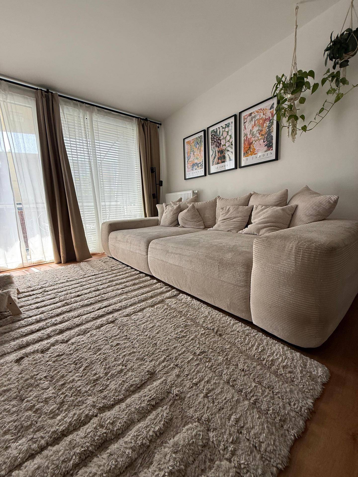 Geräumiges Wohnzimmer mit großem beigem Ecksofa, Teppich und bodentiefen Fenstern mit Vorhängen.