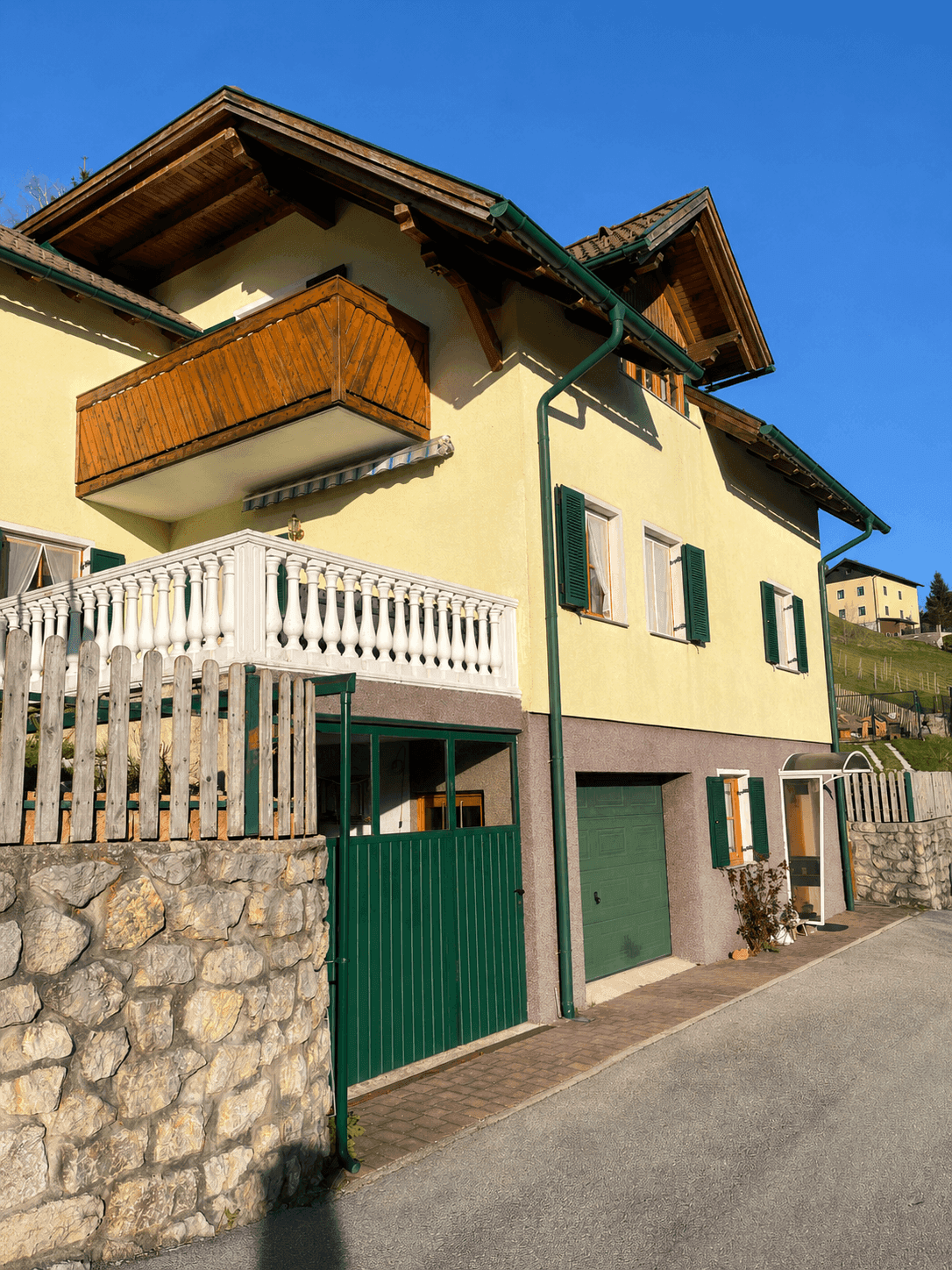 Gelbes Haus mit Holzbalkon, Steinmauer und grünen Fensterläden an einer Straße.