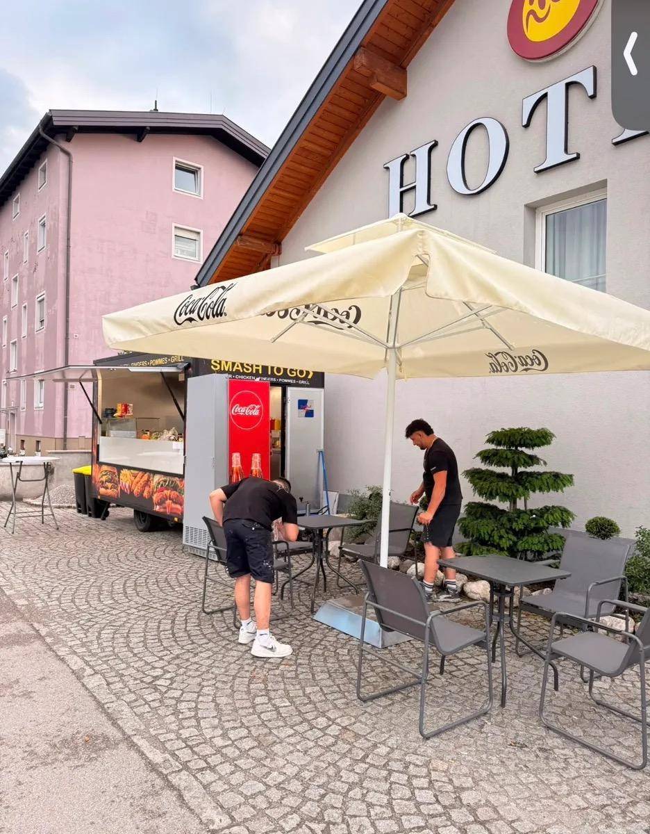Außenbereich eines Hotels mit Imbisswagen und Mitarbeitern, die Sitzgelegenheiten unter einem großen Sonnenschirm vorbereiten.