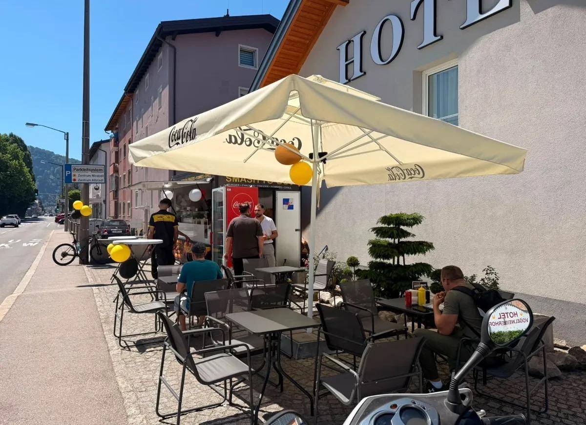 Belebtes Außengelände eines Hotels mit Imbisswagen und Gästen, die an Tischen unter einem Sonnenschirm sitzen.
