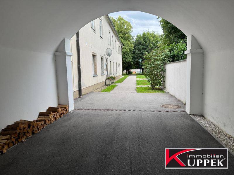 Durchgang zu einem Innenhof mit gestapeltem Brennholz und grüner Umgebung.