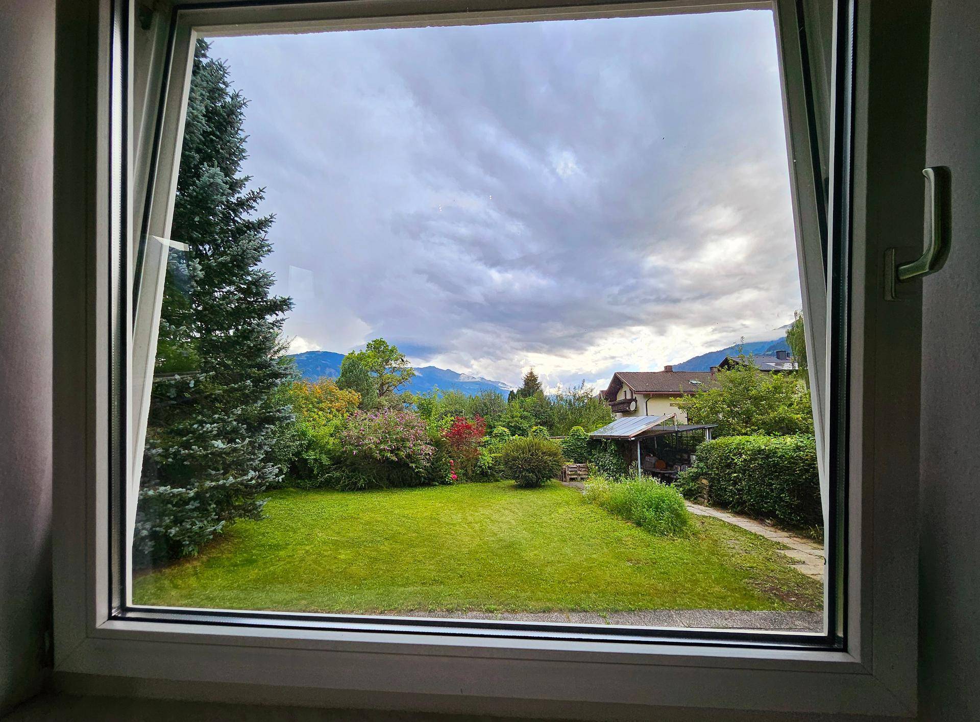 Blick aus dem Fenster auf den gepflegten Garten mit Bäumen und einer malerischen Bergkulisse im Hintergrund.