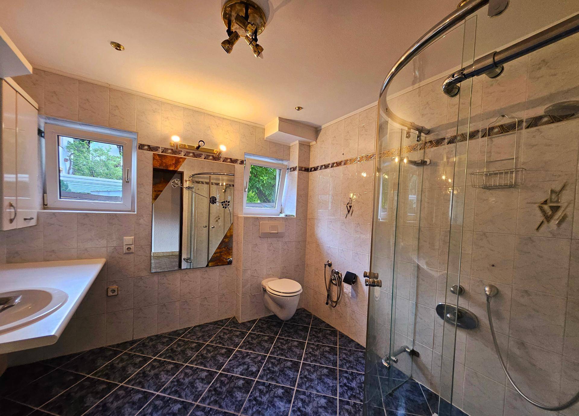 Helles Badezimmer mit Dusche, WC, Einzelwaschbecken und zwei Fenstern für Tageslicht.