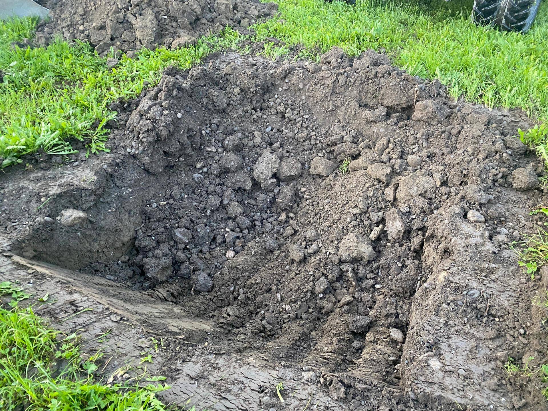 Ein Haufen ausgehobener Erde und Steine liegt neben einem kleinen Graben auf einer grünen Wiese.