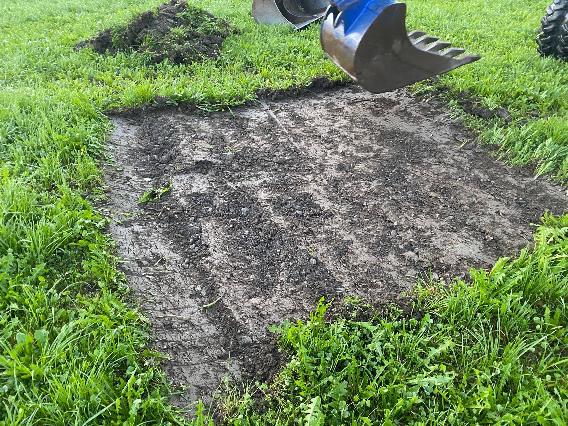 Ein Baggerlöffel schwebt über einer frisch bearbeiteten Erdfläche inmitten von Gras.
