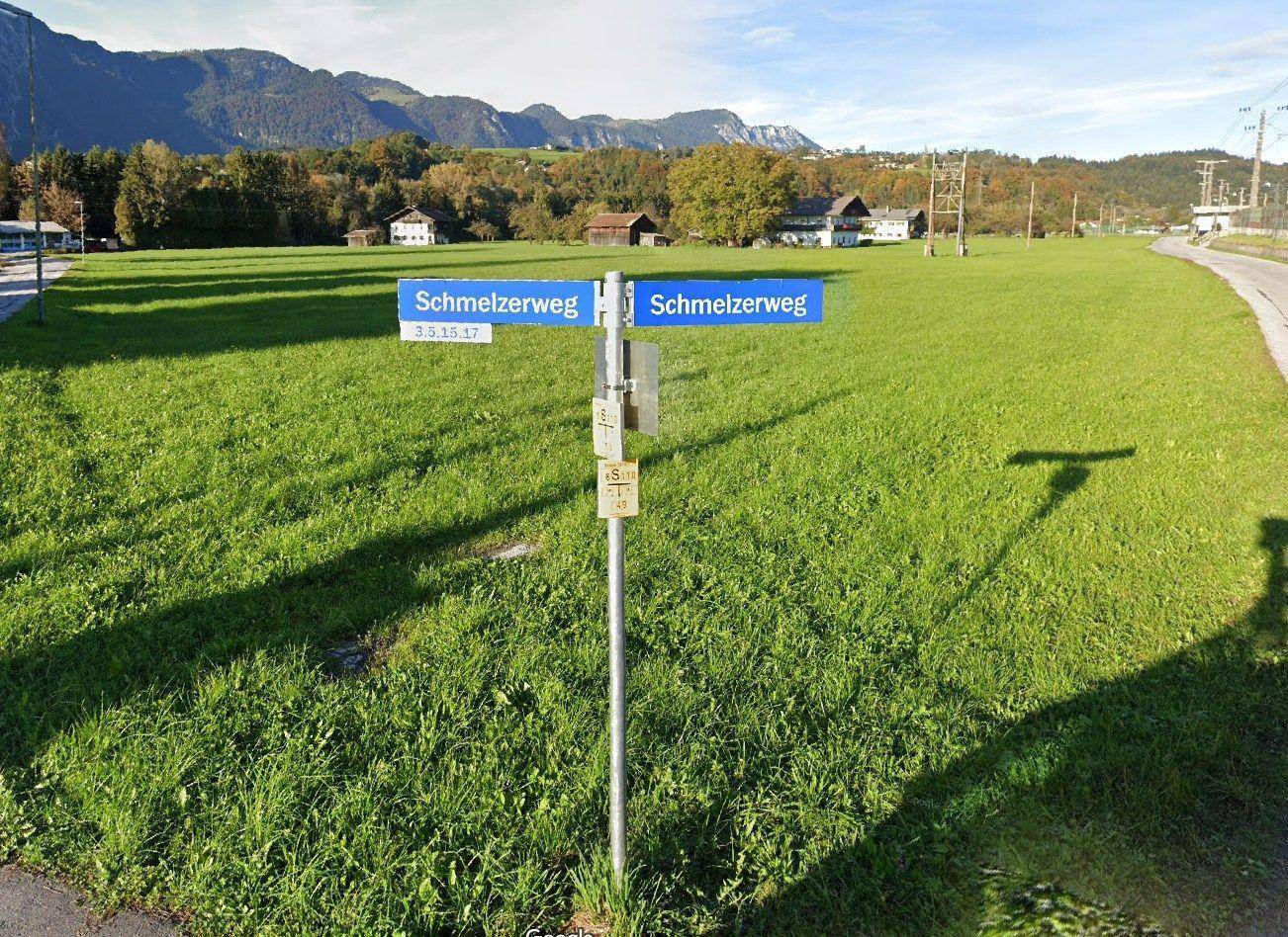 Blick auf eine grüne Wiese mit einem Straßenschild 'Schmelzerweg' und Bergen im Hintergrund.