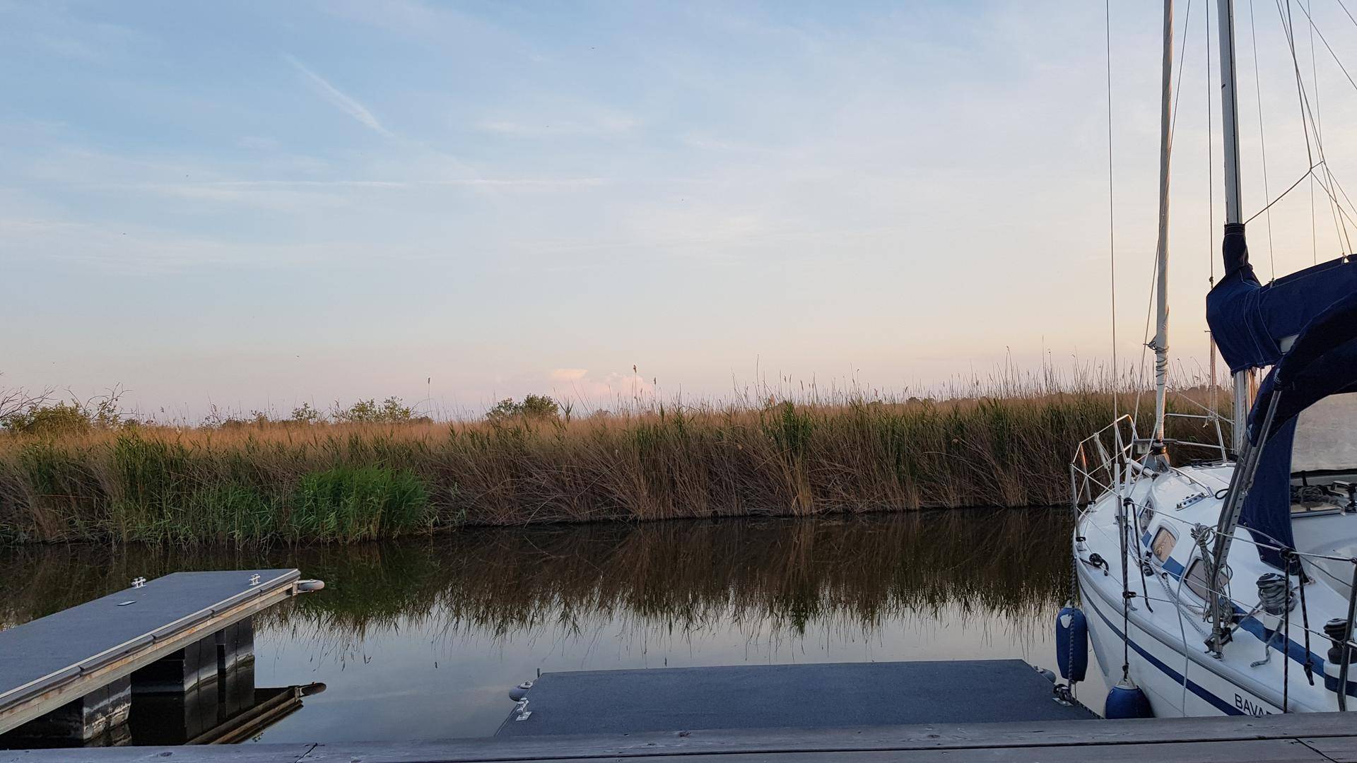 Segelboot und Anlegestelle an einem ruhigen Kanal, umgeben von Schilf.