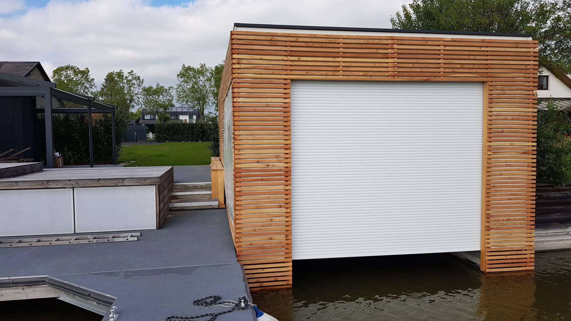 Modernes Bootshaus mit Holzfassade und großem weißen Rolltor am Wasser.