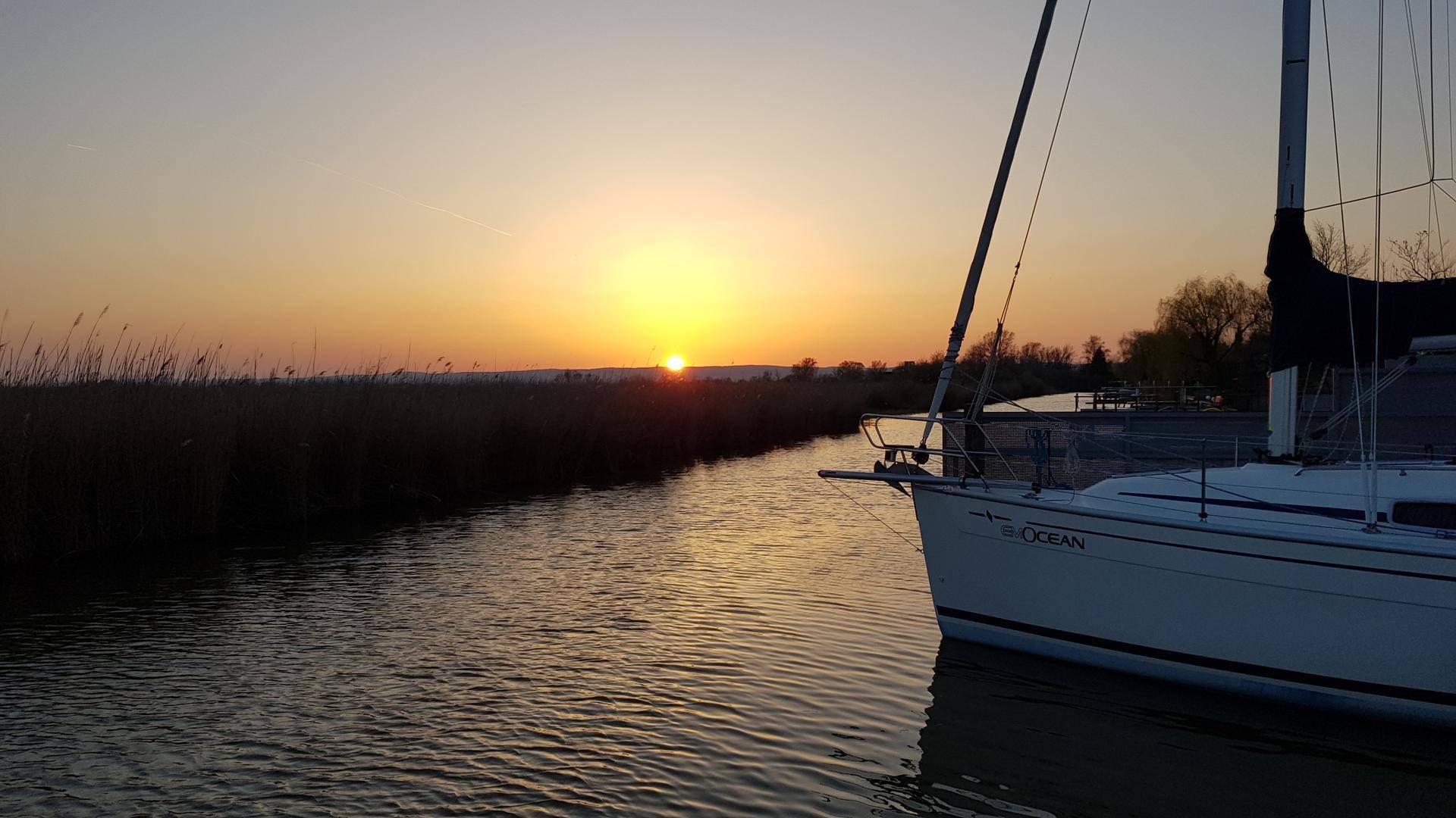 Segelboot auf dem Wasser bei Sonnenuntergang, umgeben von Schilf am Ufer.