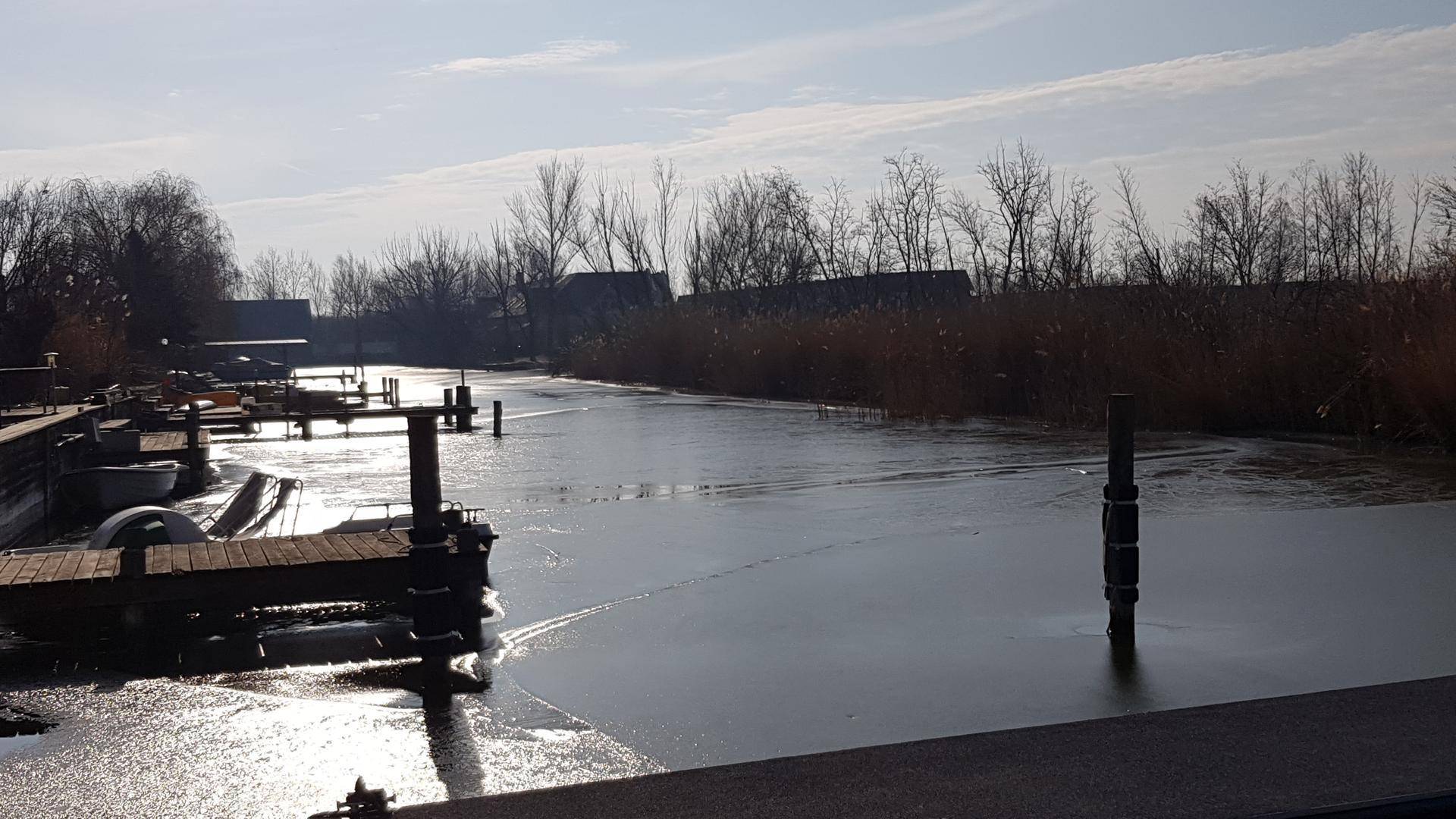 Gefrorener Kanal mit Anlegestellen und kahlen Bäumen in winterlicher Landschaft.
