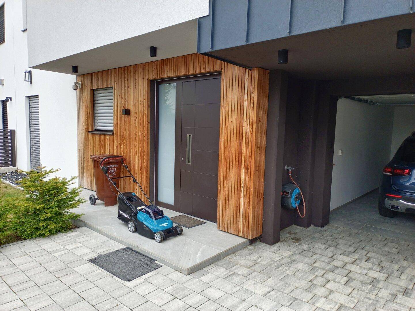 Eingangsbereich des Hauses mit Holzverkleidung, moderner Tür und angrenzendem Carport.