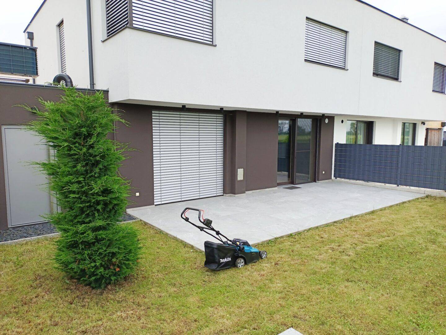 Hinterhof mit großer gepflasterter Terrasse, Rasenfläche und einem kleinen Baum.