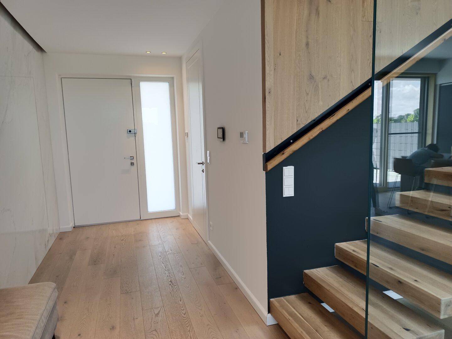 Heller Eingangsbereich mit Holzboden, weißer Tür und moderner Holztreppe mit Glasgeländer.
