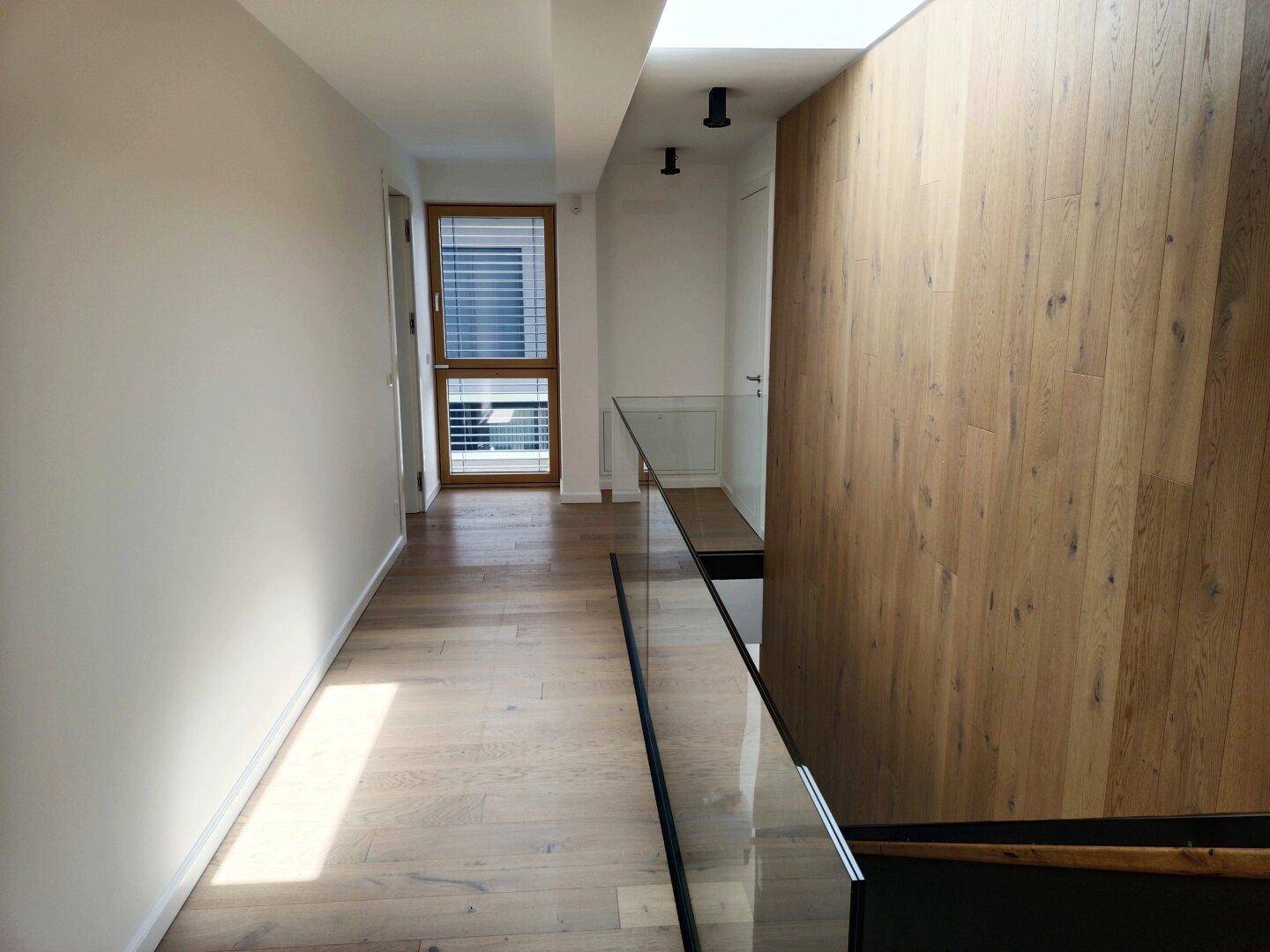 Langer, heller Flur im Obergeschoss mit Holzboden und Fenster mit Jalousien.