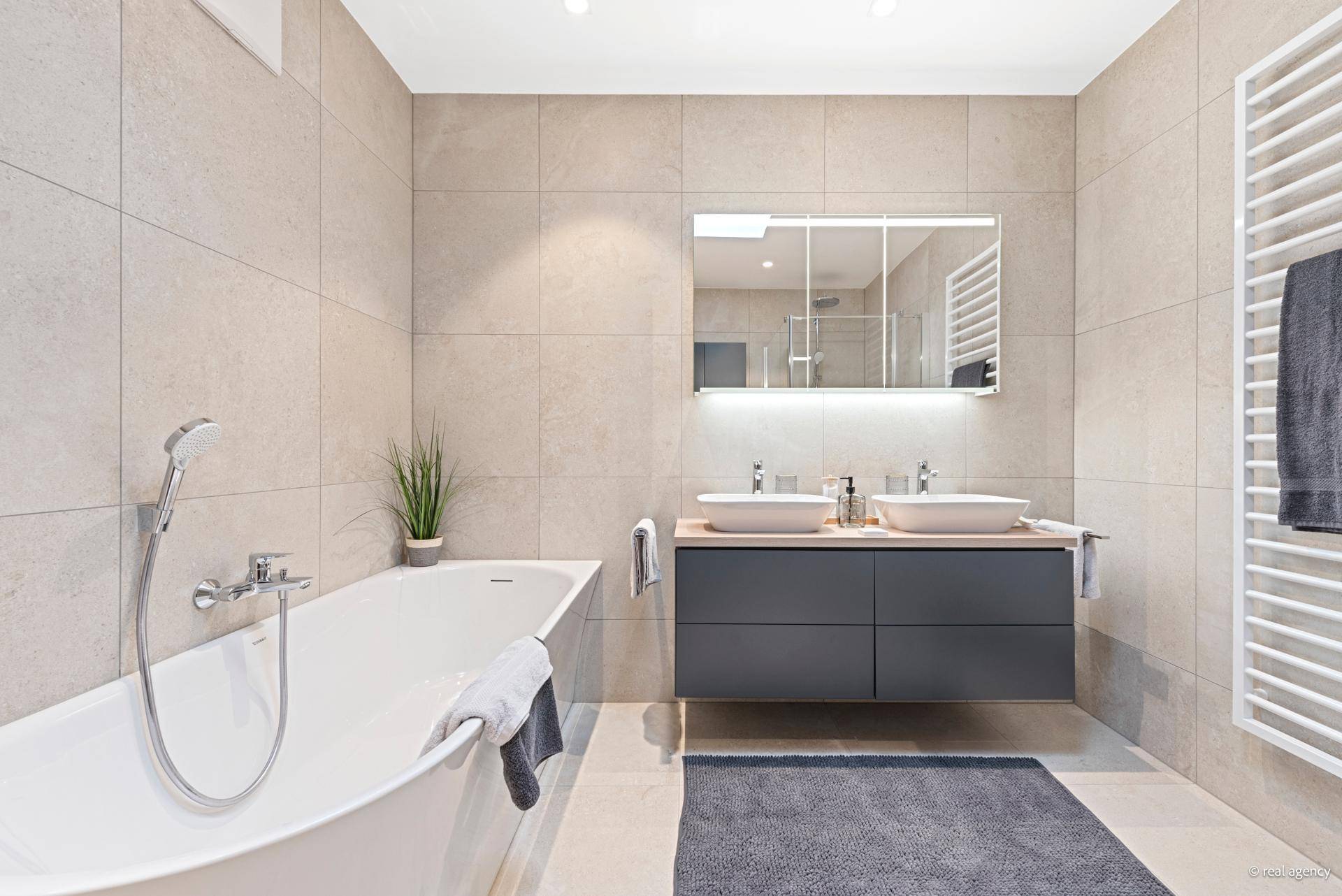 Modernes Badezimmer mit Badewanne, Doppelwaschbecken und beheiztem Handtuchhalter in hellen Farbtönen.