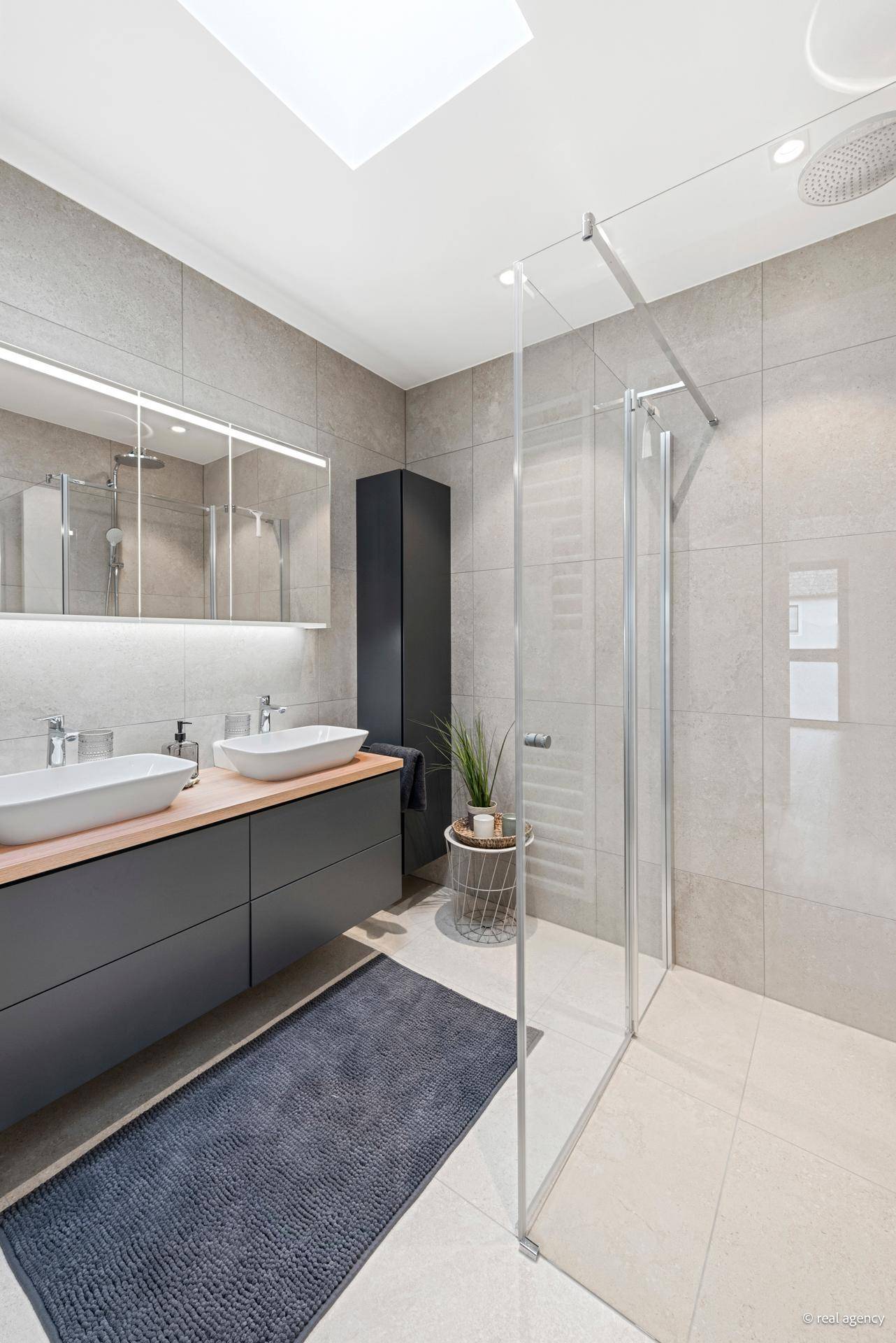 Helles Badezimmer mit Doppelwaschbecken, großer Dusche und natürlichem Licht durch ein Dachfenster.