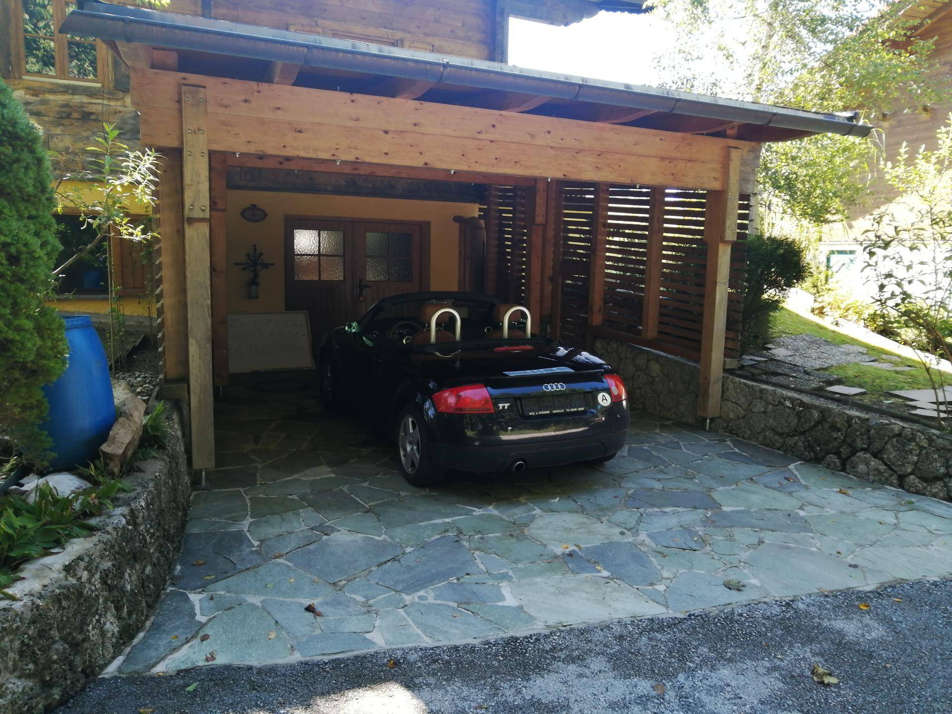 Überdachter Holzcarport mit gepflastertem Boden und einem geparkten Auto darunter.