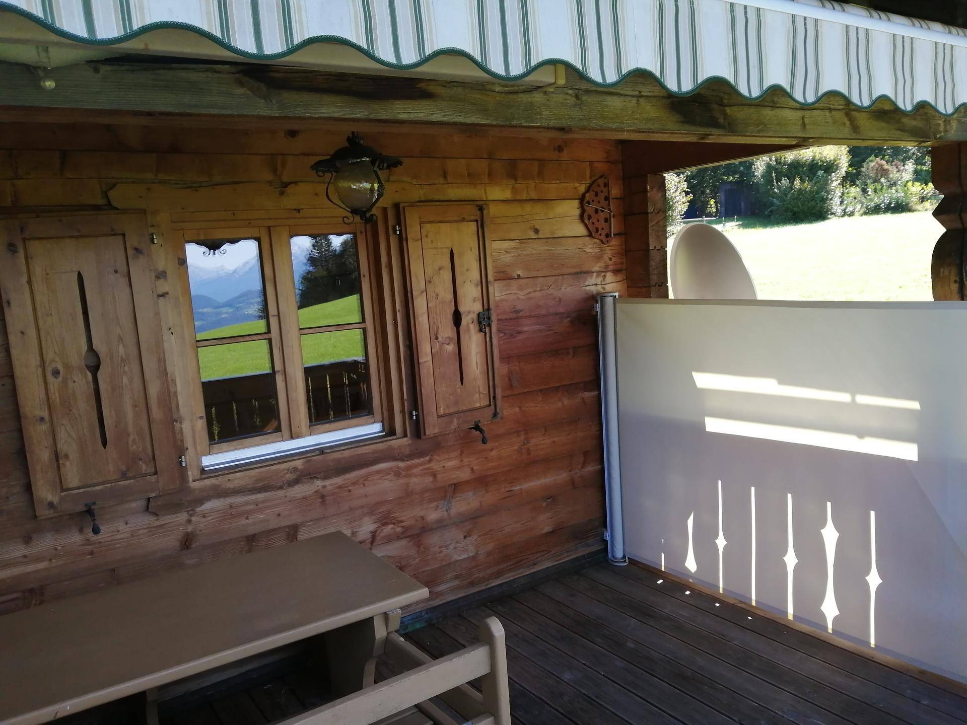 Überdachter Holzbalkon mit Sitzbank, traditionellen Fensterläden und Bergblick im Hintergrund.