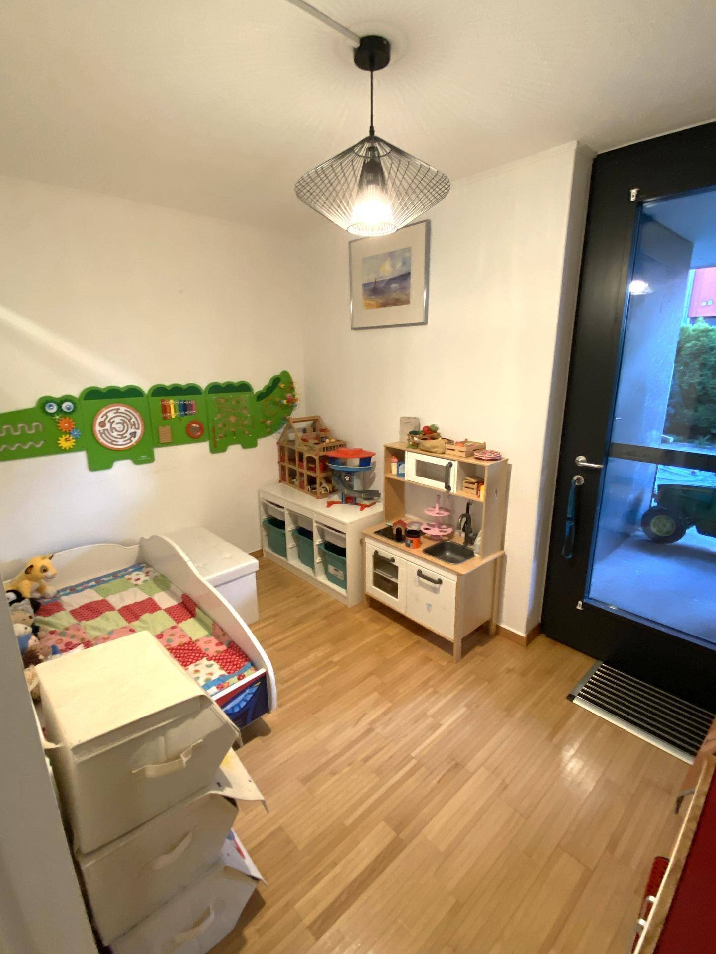 Helles Kinderzimmer mit Spielküche, Holzboden und einer großen Glastür zum Außenbereich.