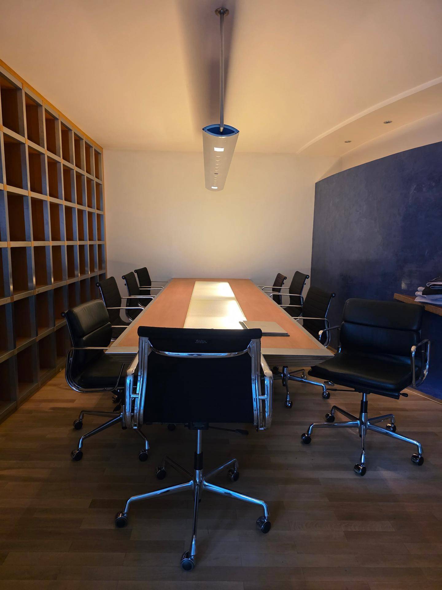 Konferenzraum mit großem Holztisch, schwarzen Bürostühlen und Holzregal an der Wand.