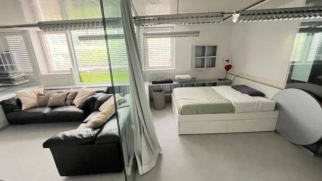 Offenes Schlafzimmer mit Bett und angrenzendem Wohnbereich mit schwarzem Ledersofa und großen Fenstern.