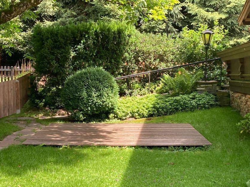Kleine Holzterrasse umgeben von gepflegtem Rasen und üppiger Bepflanzung im Garten.