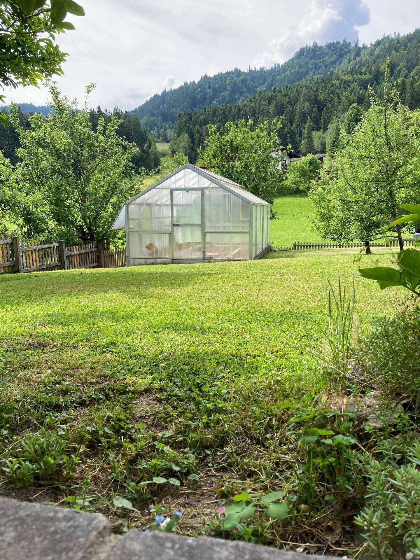 Gewächshaus in einem weitläufigen Garten mit Blick auf bewaldete Hügel und grüne Wiesen.