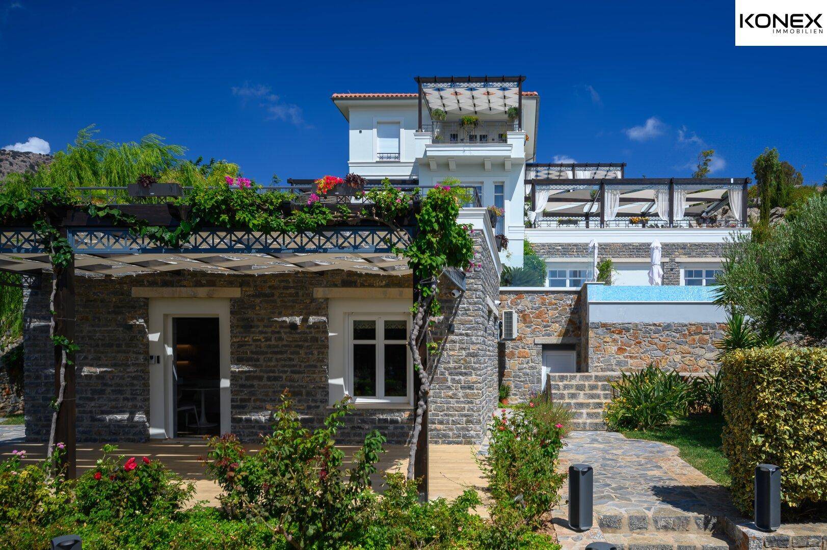 Steinverkleidete Fassade der Villa mit üppiger Bepflanzung und Treppen, die zum Garten führen.