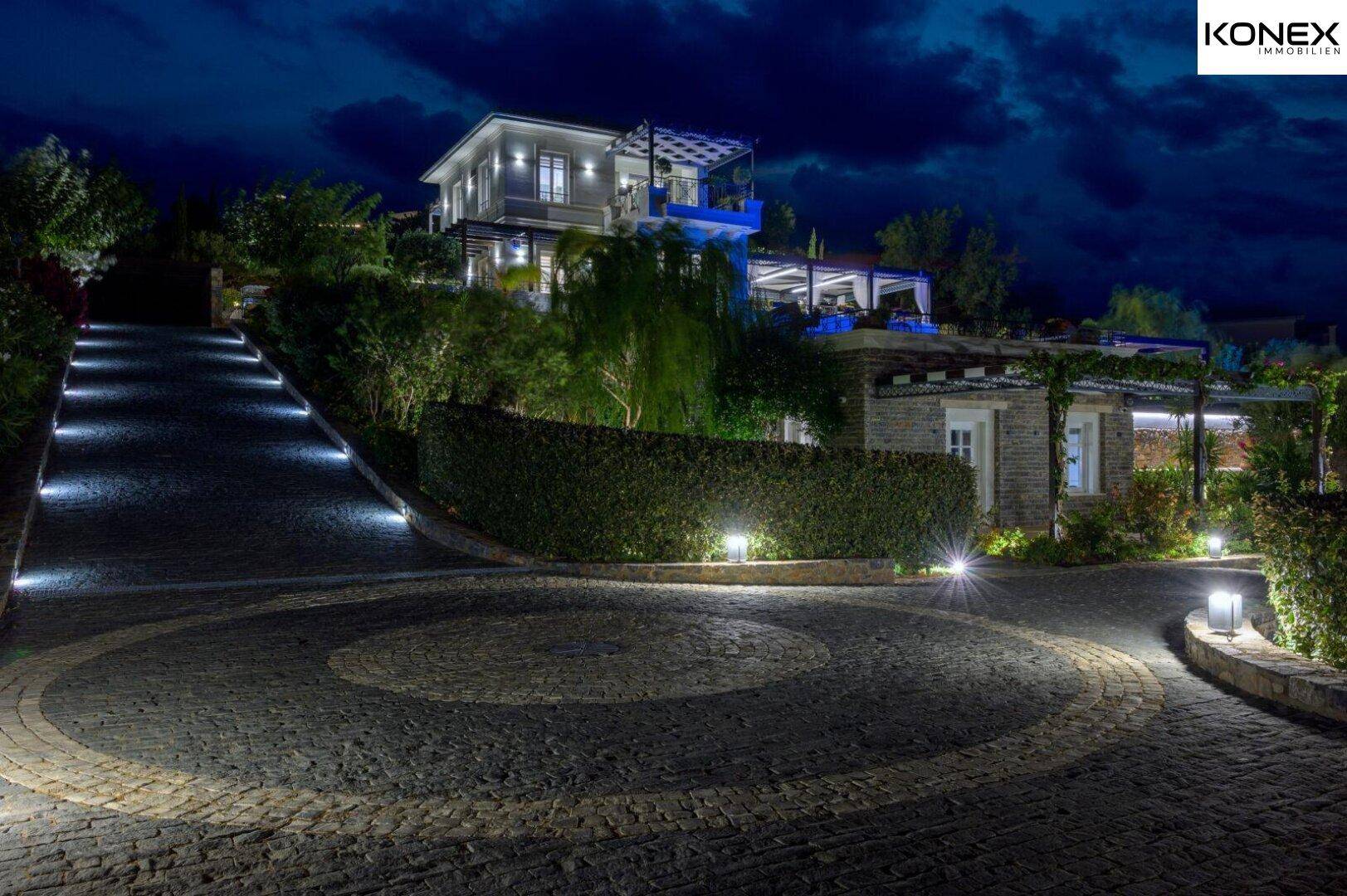 Beleuchtete Auffahrt zur Villa bei Nacht, umgeben von gepflegten Hecken und Bäumen.