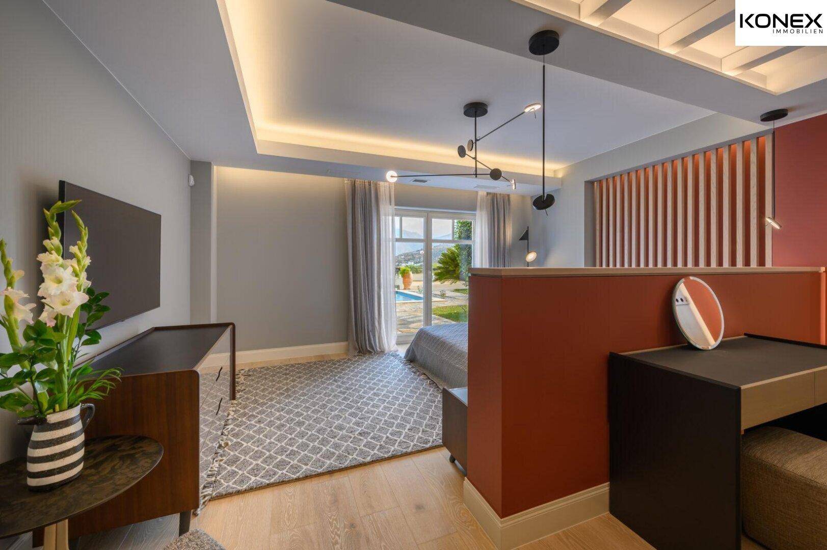 Modernes Schlafzimmer mit Holzboden, roter Akzentwand und Blick auf den Poolbereich.