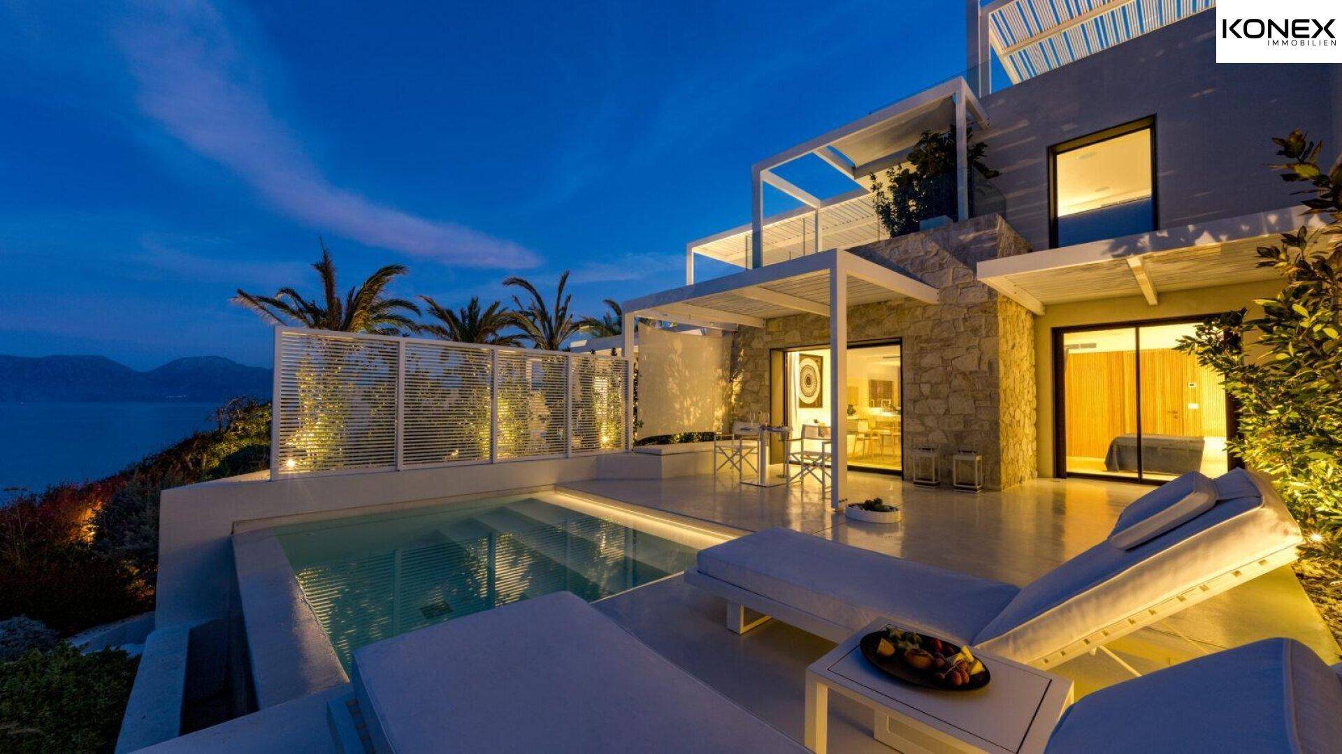 Luxuriöse private Terrasse mit Pool und Liegen bei Dämmerung, mit Blick auf das Meer und die beleuchtete Villa.