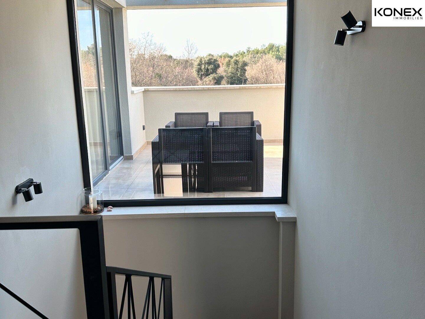 Treppenaufgang mit Blick auf eine möblierte Dachterrasse durch große Fenster.
