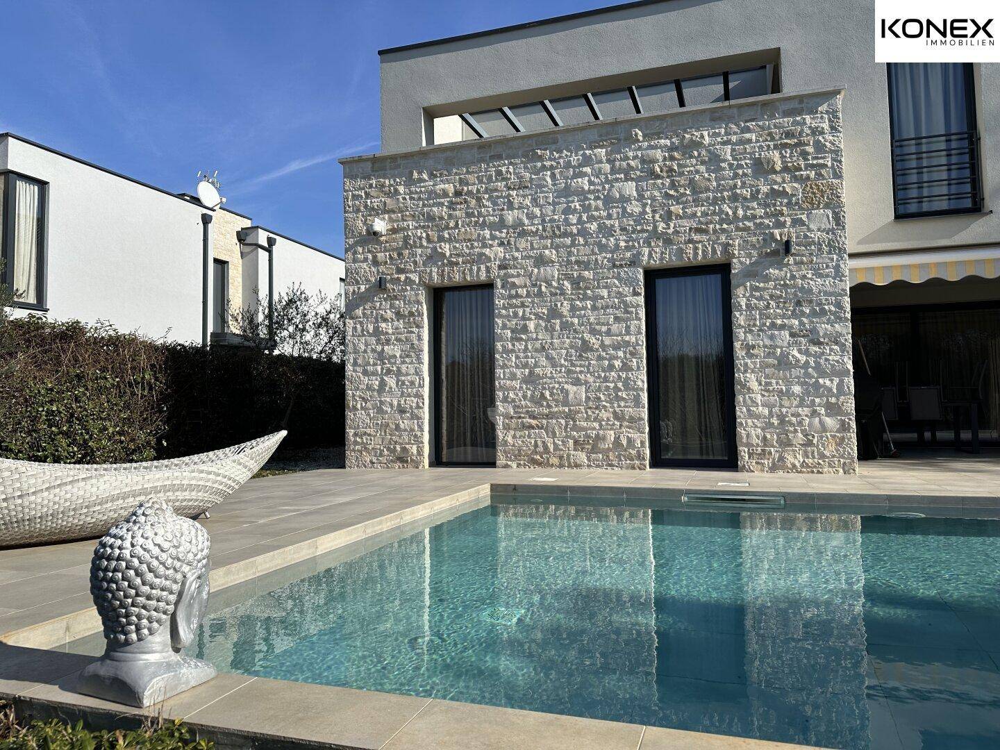 Moderne Villa mit Steinfassade und großem Swimmingpool im Außenbereich an einem sonnigen Tag.